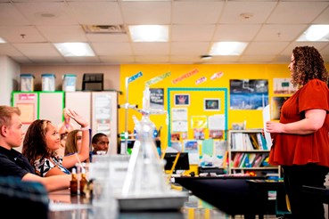 A high school science teacher speaks to a room full of students. Some students have their hands raised to speak, and there is chemistry equipment in the foreground on a desk.