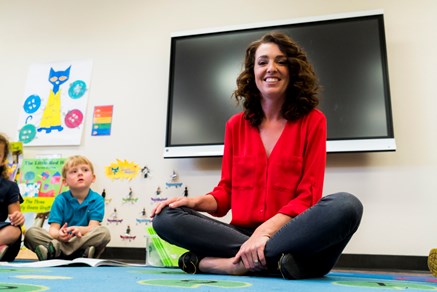 A teacher sits crosslegged on a colorful rug in her classroom as she smiles at the camera.